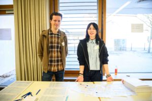 Zwei Personen stehen hinter einem Tisch mit Anmeldebögen. Eine Person lehnt sich mit beiden Händen gegen die Tischkante.