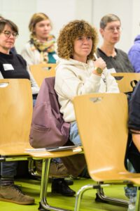 Mehrere Personen sitzen in Stuhlreihen und schauen nach vorne, einige der Stühle sind leer.