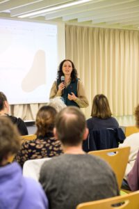 Auf einer Bühne befindet sich im Hintergrund eine Leindwand, auf die etwas projeziert wird. Vor der Bühne steht eine Person mit langen Haaren und Mikrofon in der einen Hand, während sie mit der anderen Hand gestikuliert. Sie spricht dabei. Vor der Bühne sitzen auf Stühlen Personen die zu der Person auf der Bühne blicken und zuhören.