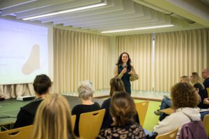 Auf einer Bühne befindet sich im Hintergrund eine Leindwand, auf die etwas projeziert wird. Davor steht eine Person mit langen Haaren und Mikrofon in der einen Hand, während sie mit der anderen Hand gestikuliert. Sie spricht dabei. Vor der Bühne sitzen auf Stühlen Personen die zu der Person auf der Bühne blicken und zuhören.