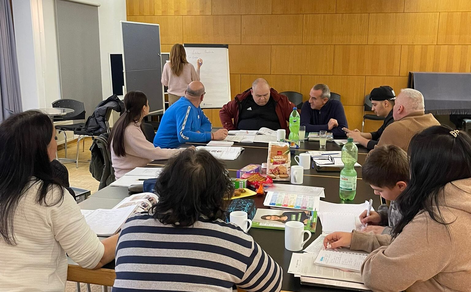 An einem großen Tischblock sitzen 10 Personen, sie haben Schreibutensilien vor sich auf dem Tisch ausgebreitet. Im Hintergrund ist eine Person, die etwas an ein Flipchart schreibt.