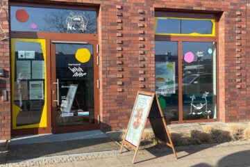 Bunt gestaltete Schaufenster und die Eingangstür des Quartierstreffs Jetzt Hier. Vor dem Eingang steht ein Aufsteller mit einem Plakat.