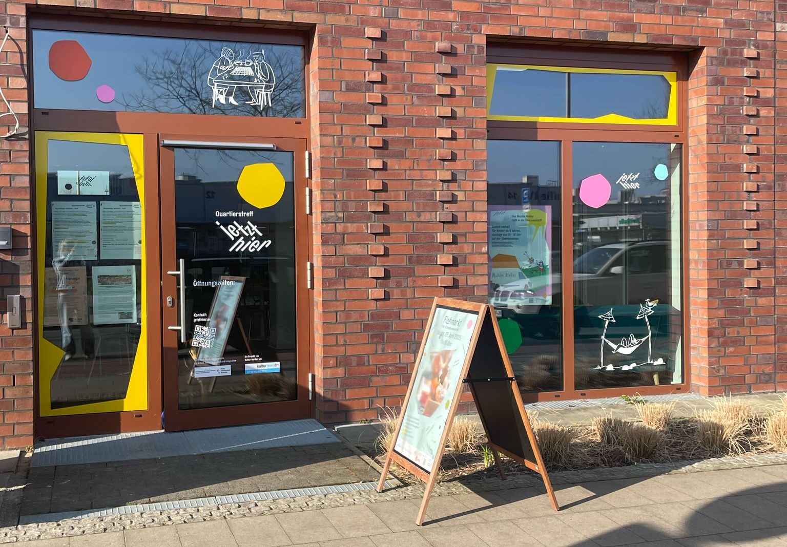 Bunt gestaltete Schaufenster und die Eingangstür des Quartierstreffs Jetzt Hier. Vor dem Eingang steht ein Aufsteller mit einem Plakat.